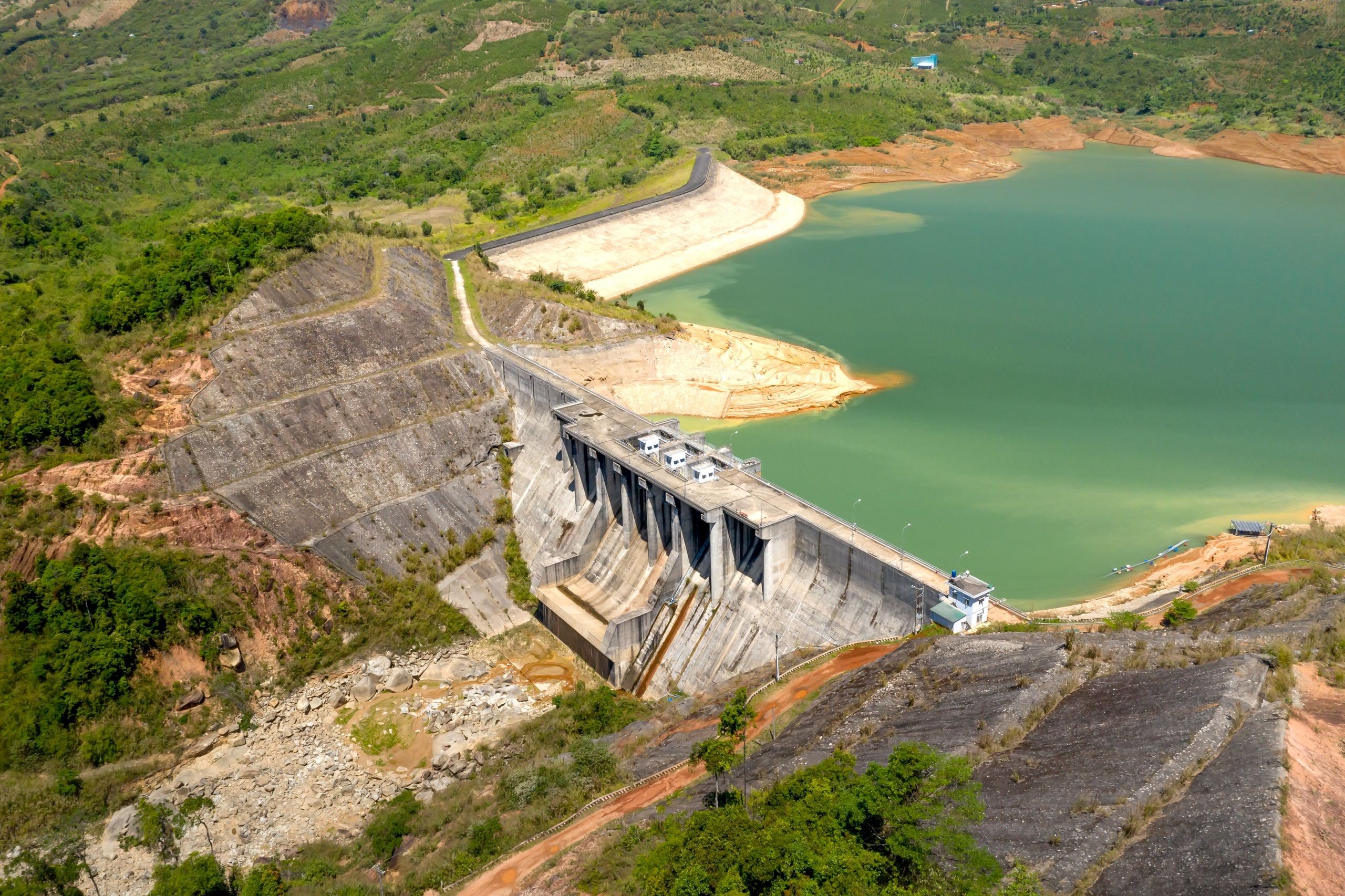 Aerial view of a concrete hydropower dam and its green reservoir, between hills of red earth
