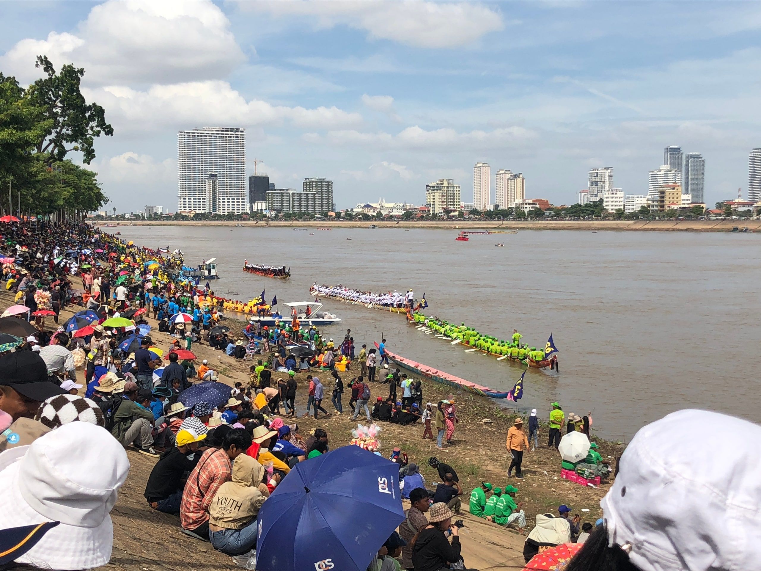 La foule sous les parapluies sur la berge du Tonlé Sap à Phnom Penh, pendant que des pirogues de course attendent au bord du fleuve