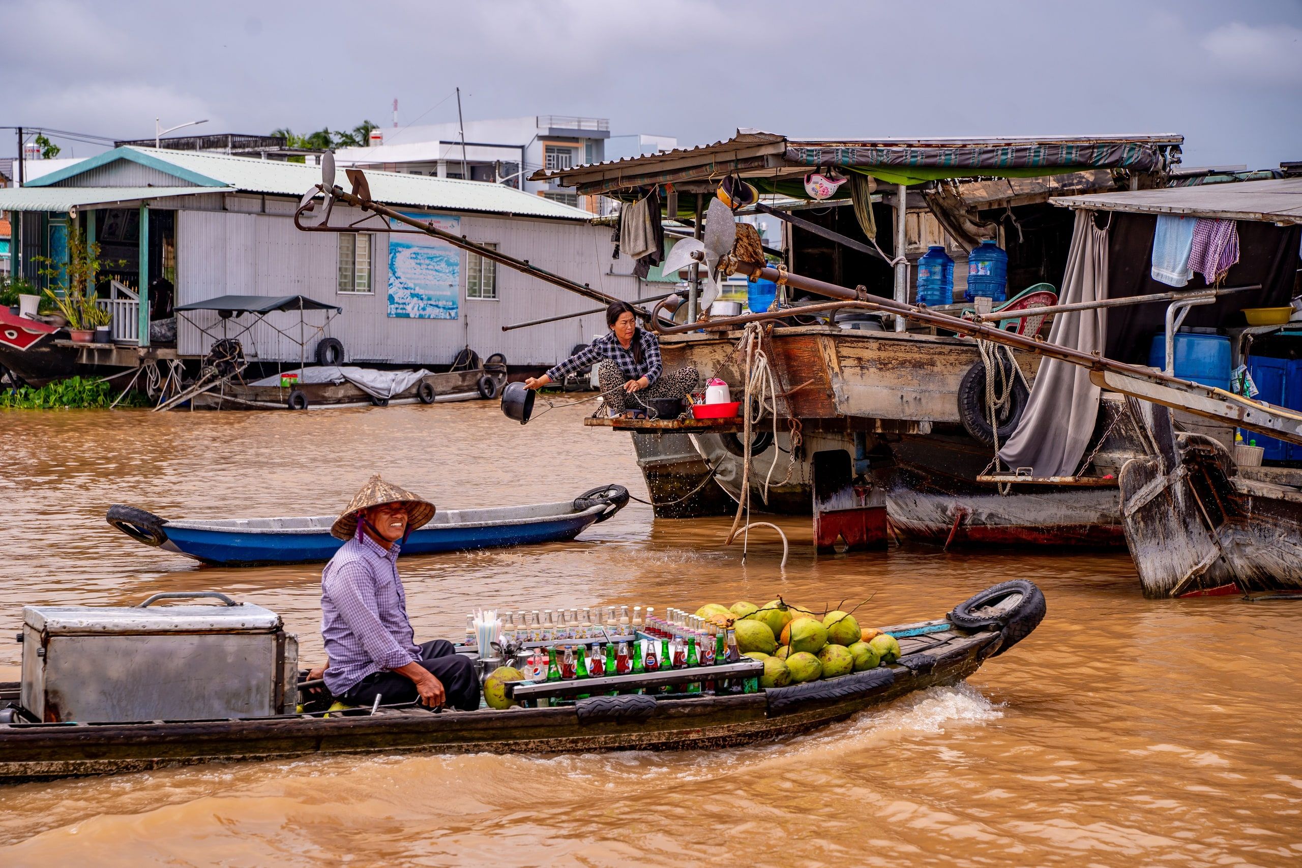 A vendor in a conical hat on his boat loaded with coconuts and drinks, in front of houseboats, at the Can Tho floating market
