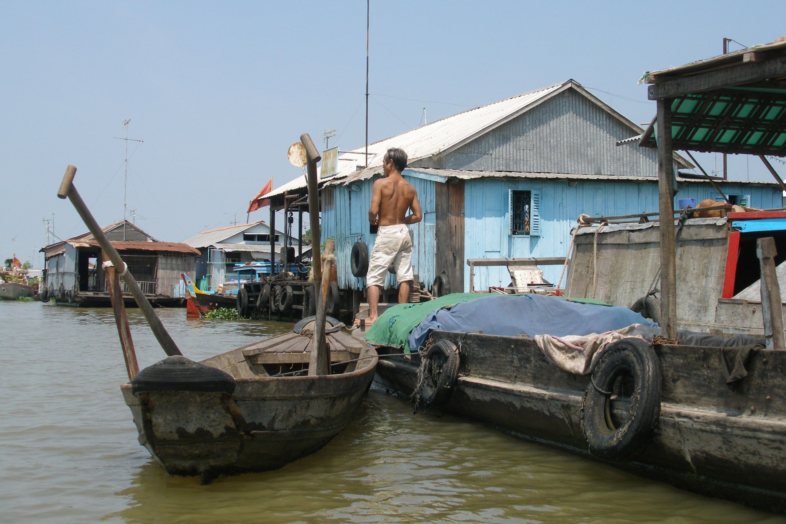 Un homme debout sur une barque devant les maisons flottantes du Bassac
