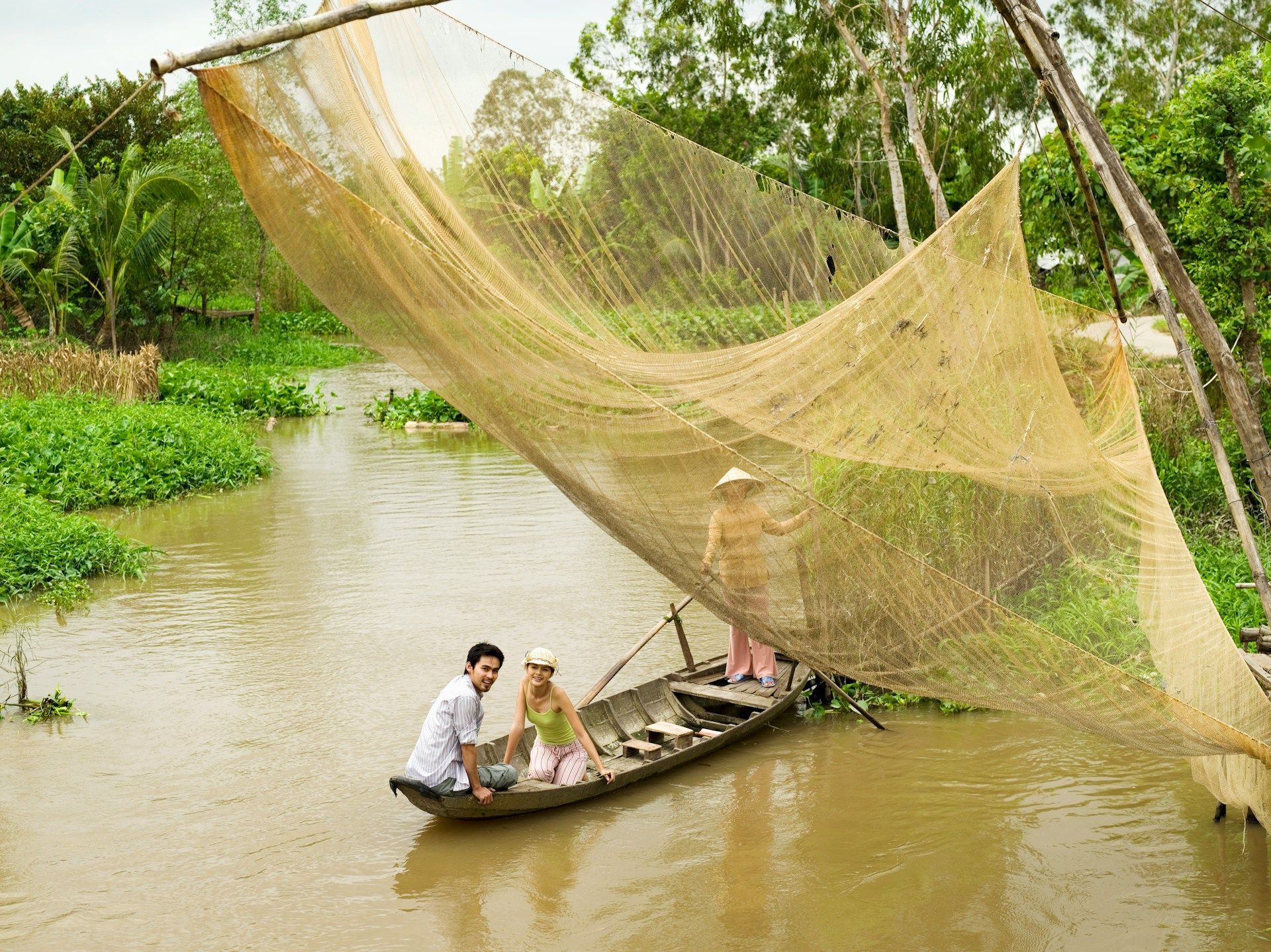 Two travelers in a rowing boat under a large fishing net raised above a delta canal