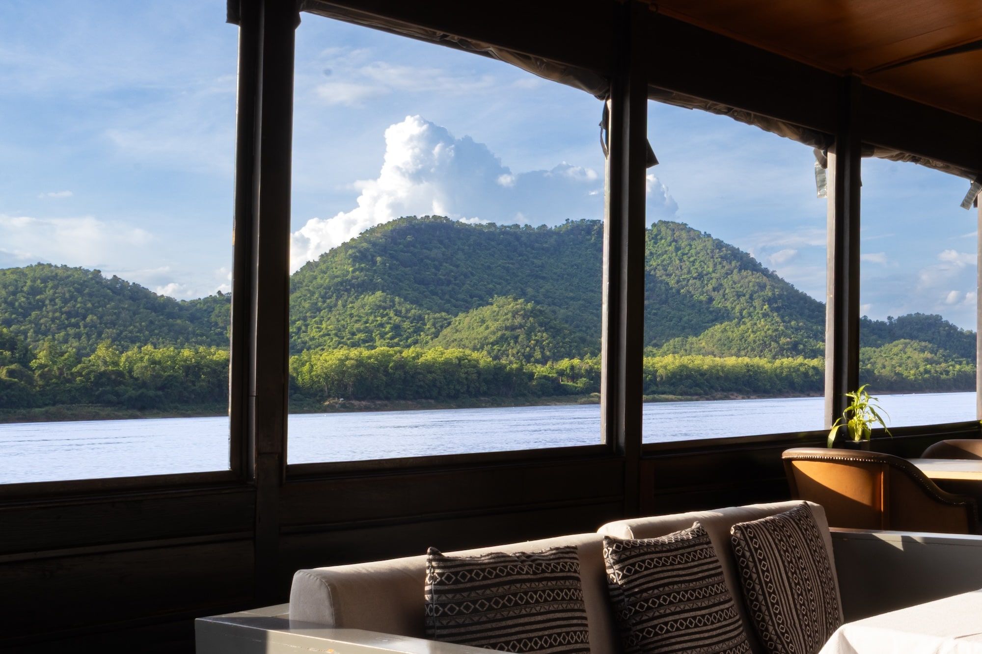 Le salon d'un bateau de croisière, banquettes et coussins tissés face au fleuve et aux collines boisées