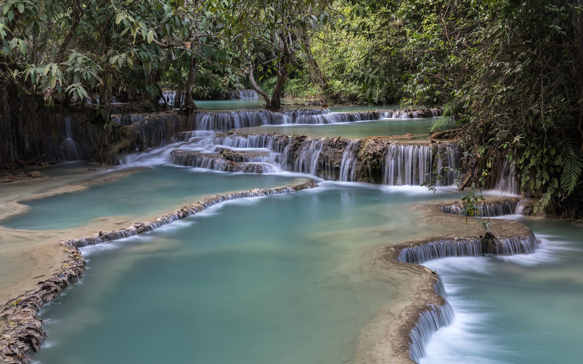 Une suite de vasques turquoise en paliers, bordées de calcaire et de forêt, en contrebas des chutes de Kuang Si