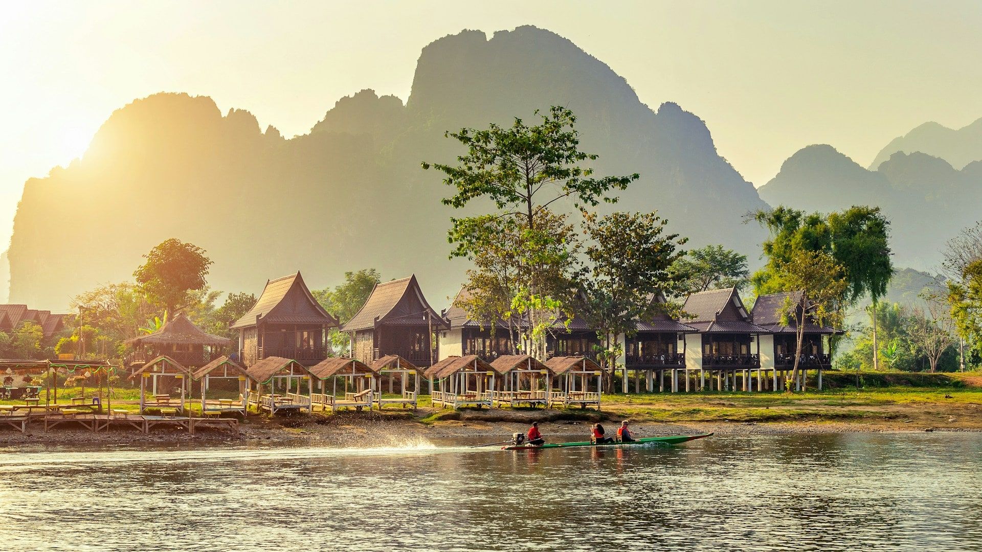 Bungalows on a riverbank below limestone peaks, in the evening light