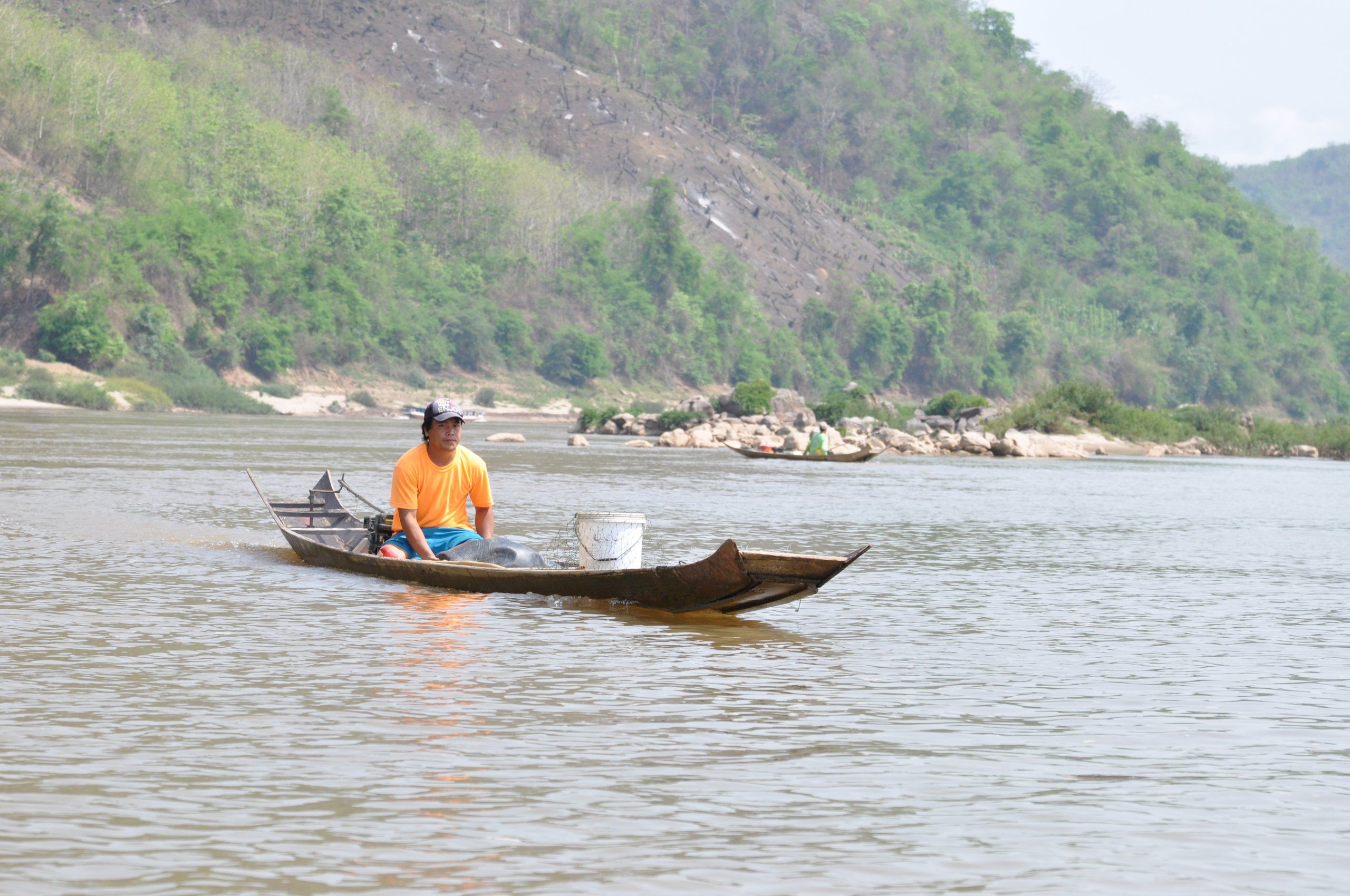 Un pêcheur en pirogue sur le Mékong, devant des rochers à découvert au pied de collines boisées