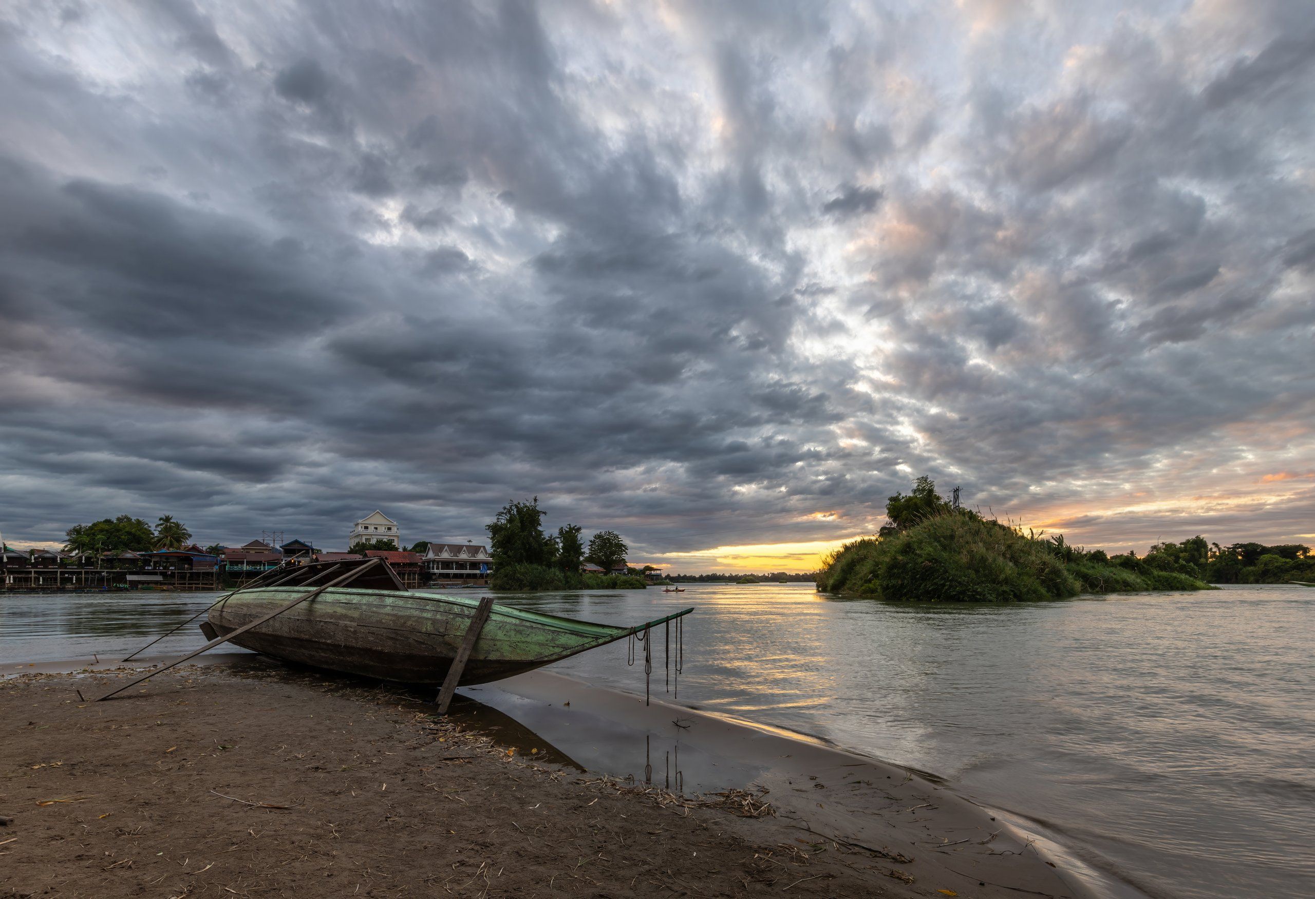 A long boat pulled up on a sandy Mekong beach in the 4,000 Islands, under a cloudy sunset sky
