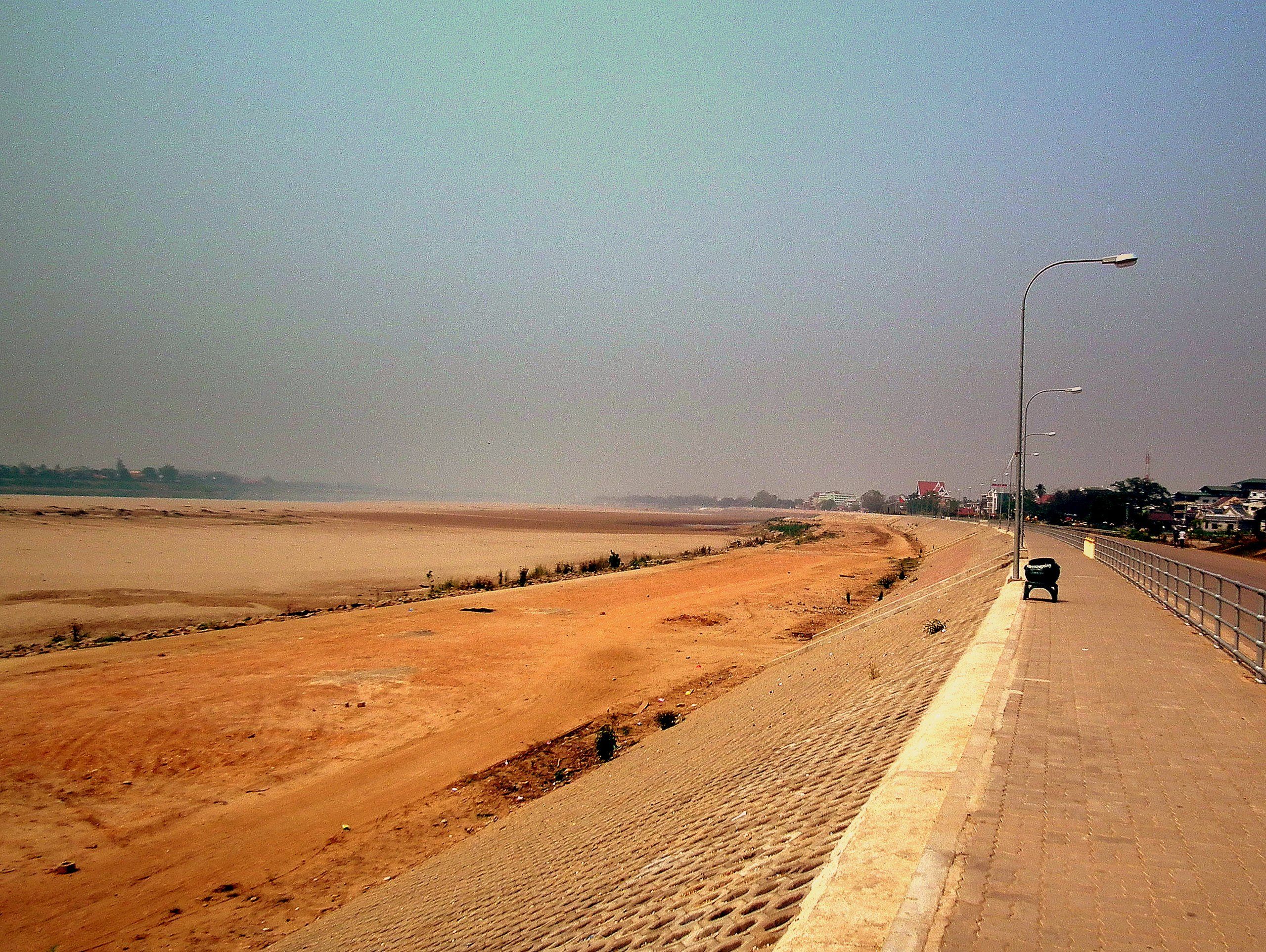 A wide sandy bank exposed along the Mekong in Vientiane, under a hazy sky, beside the embankment