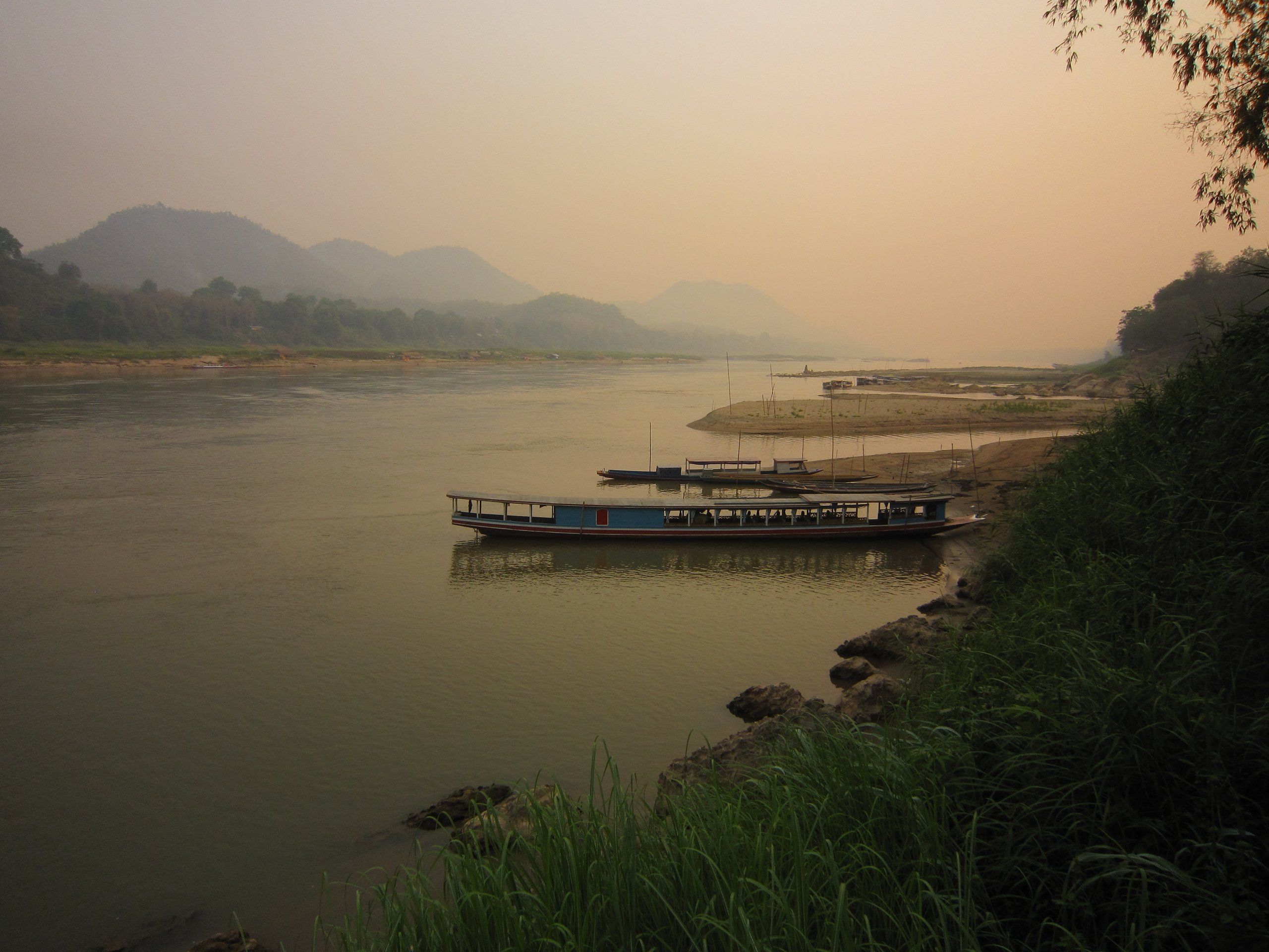 The Mekong at Luang Prabang under a hazy sky, boats moored below a wooded bank