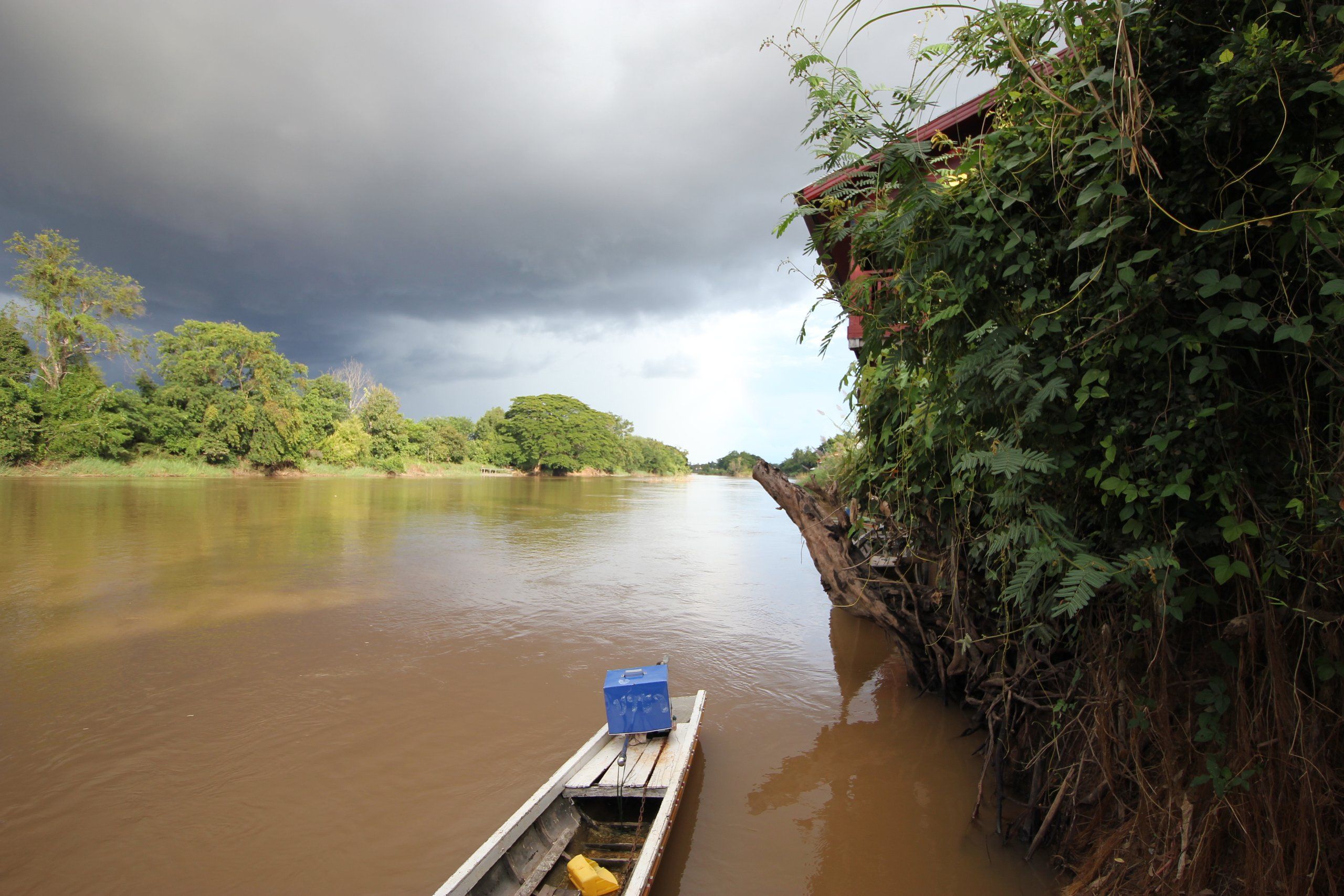 A long boat on the brown Mekong in the 4,000 Islands, under a stormy sky, between green banks