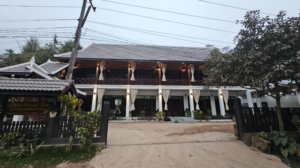 La façade du Charme Pakbeng Villa, une maison blanche à galerie de bois et rideaux clairs, au fond d'une cour de sable