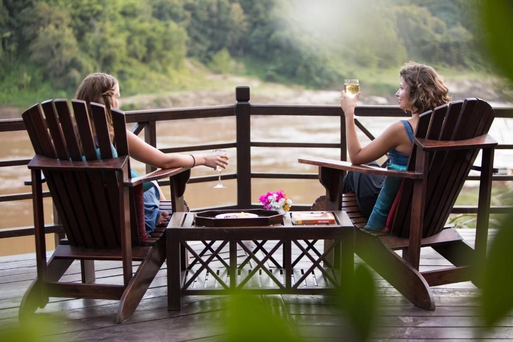 Two travelers in wooden armchairs on the terrace of Luang Say Lodge, glasses in hand, above the brown Mekong