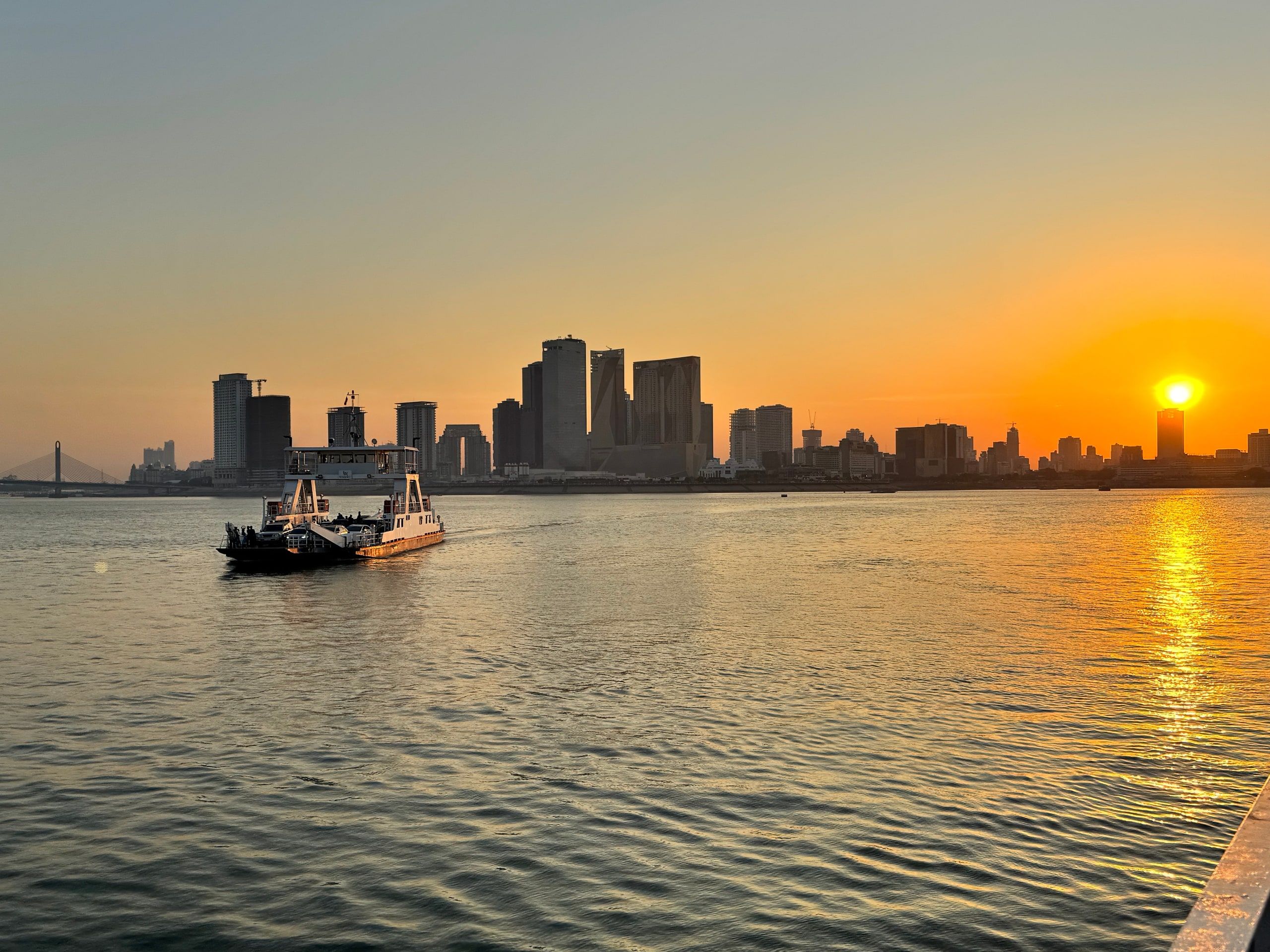 The sun sets behind the Phnom Penh skyline, a boat on the river in the foreground