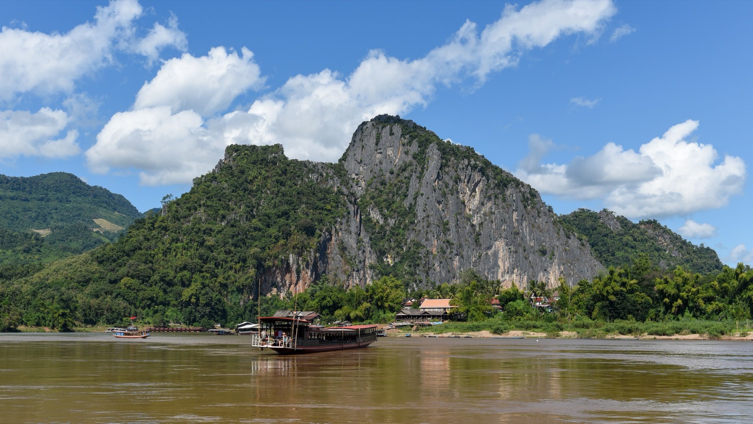A slow boat on the Mekong at the foot of a forested limestone cliff