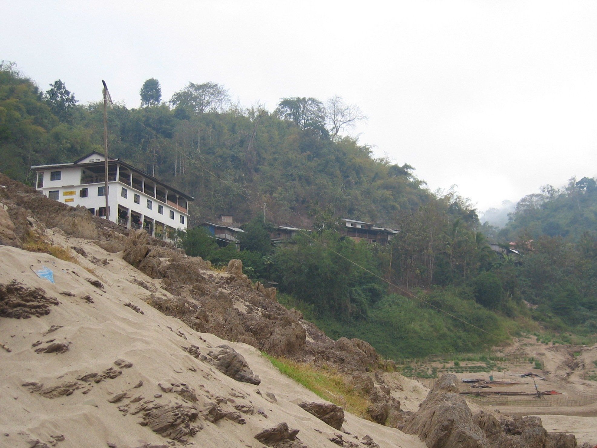 Pakbeng vu du Mékong : une maison blanche à étage et des toits sur la colline, au-dessus d'une berge de sable