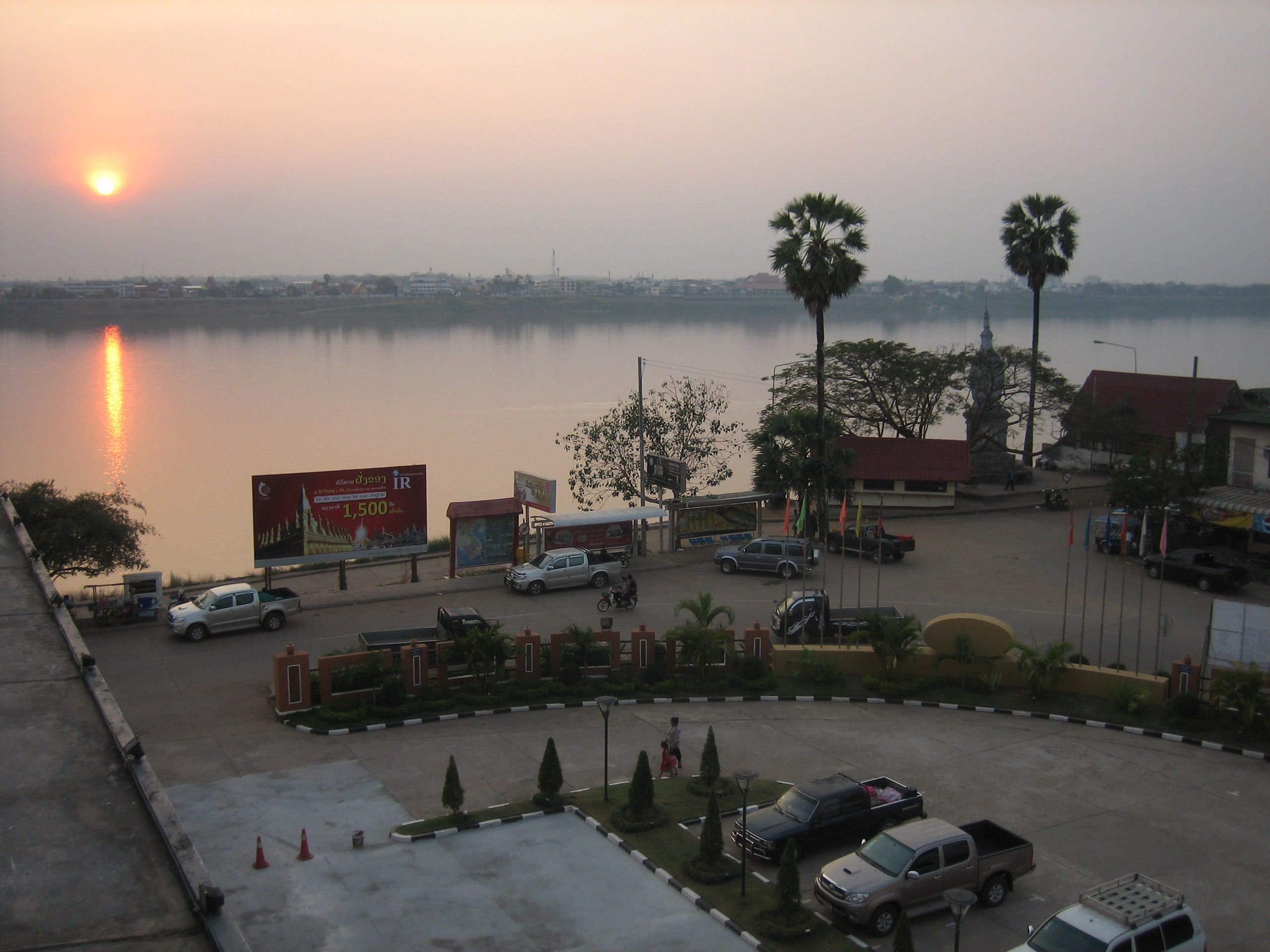 Le soleil se couche sur le Mékong vu de Thakhek, la rive thaïe à l'horizon, des palmiers et une place au bord de l'eau