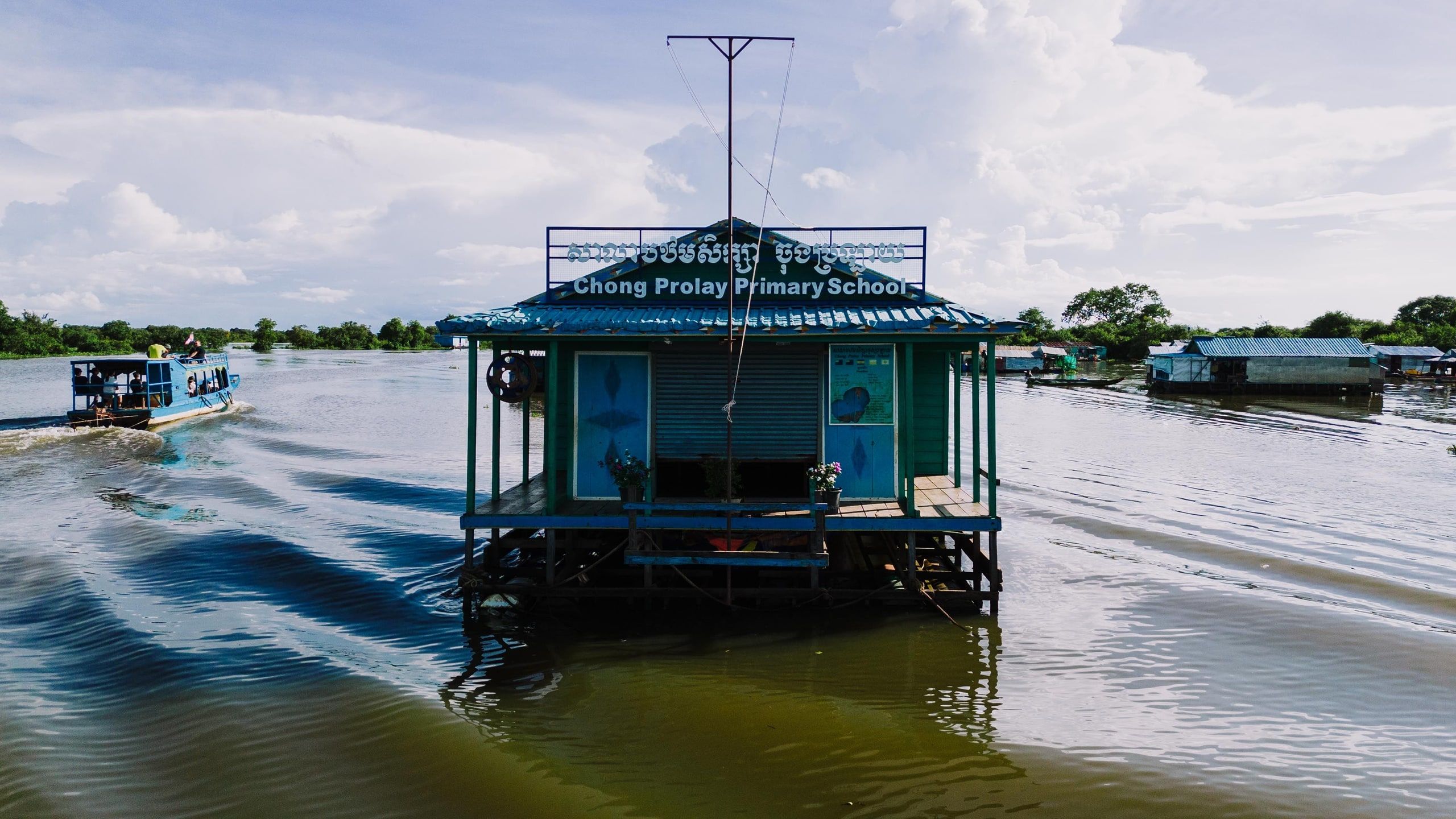 A floating school painted green and blue, with a “Chong Prolay Primary School” sign, sitting on the lake; a boat passes on the left